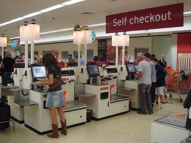 A row of self-service checkout machines in a UK supermarket, each with a touchscreen, a scanner and a card reader.